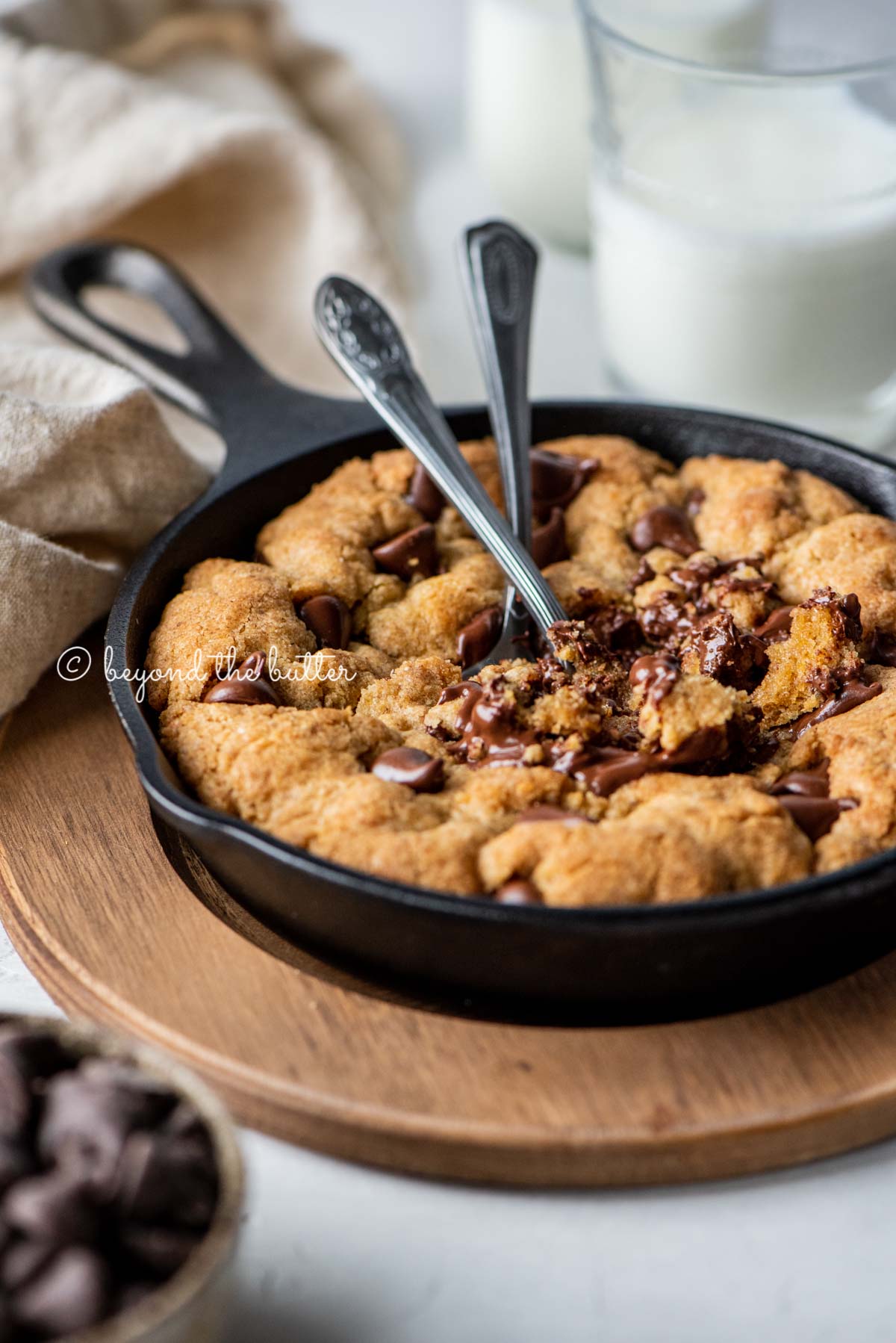 Warm brown butter chocolate chip skillet cookie with two spoons resting in skillet on wood trivet | All Images © Beyond the Butter®