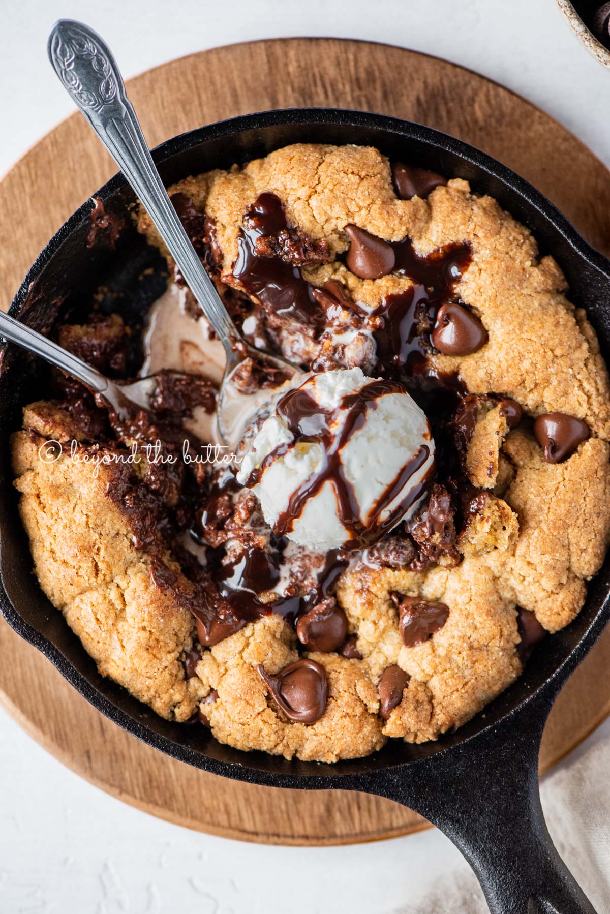 Closeup of brown butter chocolate chip skillet cookie with scoop of vanilla ice cream and two spoons resting in skillet on wood trivet | All Images © Beyond the Butter®