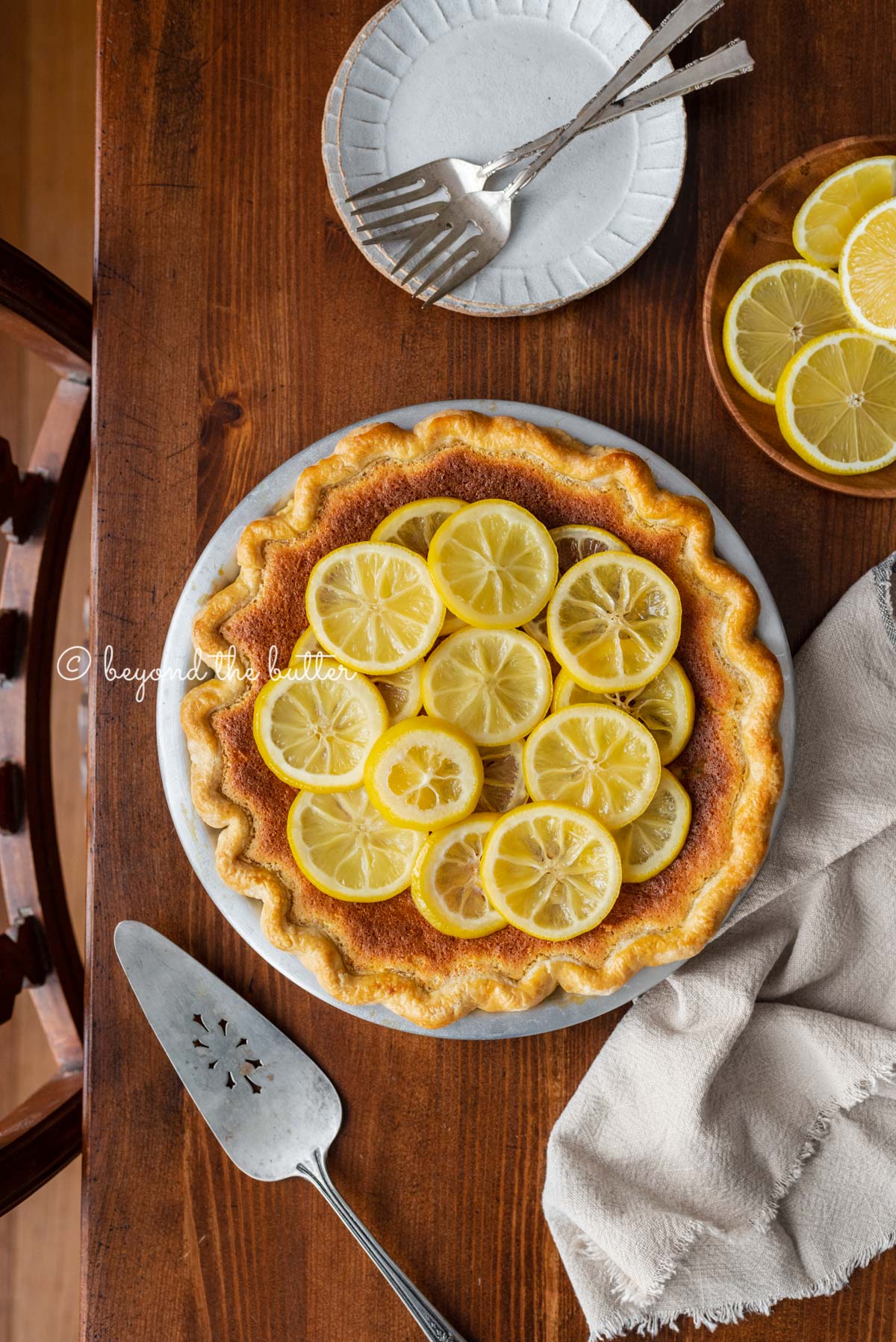 lemon sponge pie decorated with candied lemon sliced on a dark wood background with small dessert plates, napking, pie server, and small plate of sliced lemons | All images © Beyond the Butter®