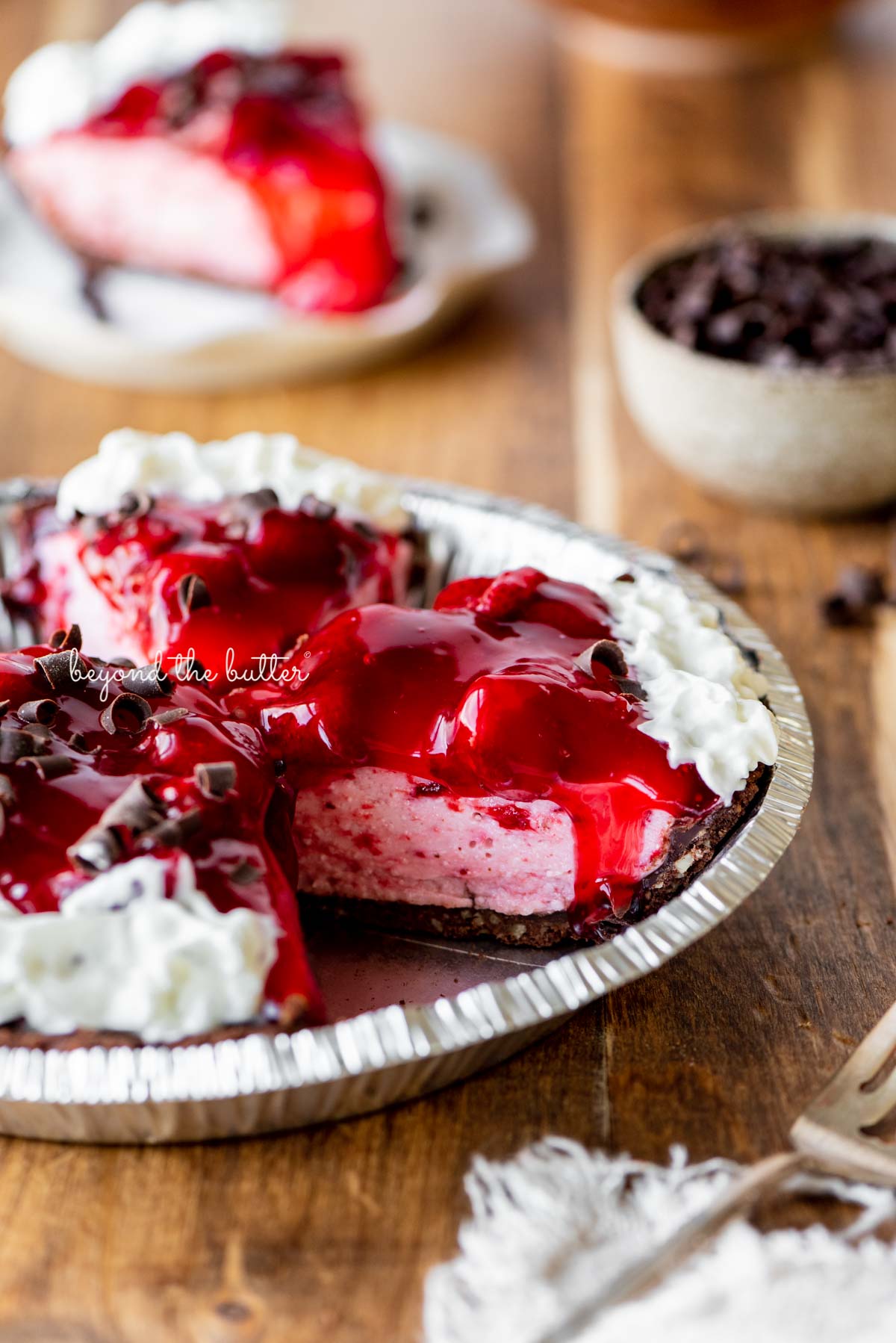 Sliced no bake strawberry chocolate cream pie on wood table with a fork and small bowl of chocolate curls nearby.