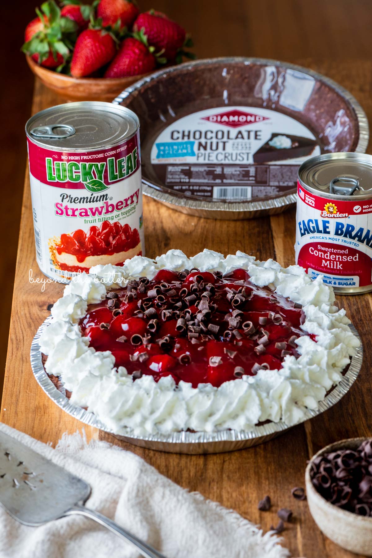 Diamond of CA® chocolate nut pie crust, Lucky Leaf® strawberry pie filling, and Eagle Brand® sweetened condensed milk with decorated no bake strawberry chocolate pie with small bowl of chocolate curls and strawberries on wood table | All images © Beyond the Butter®
