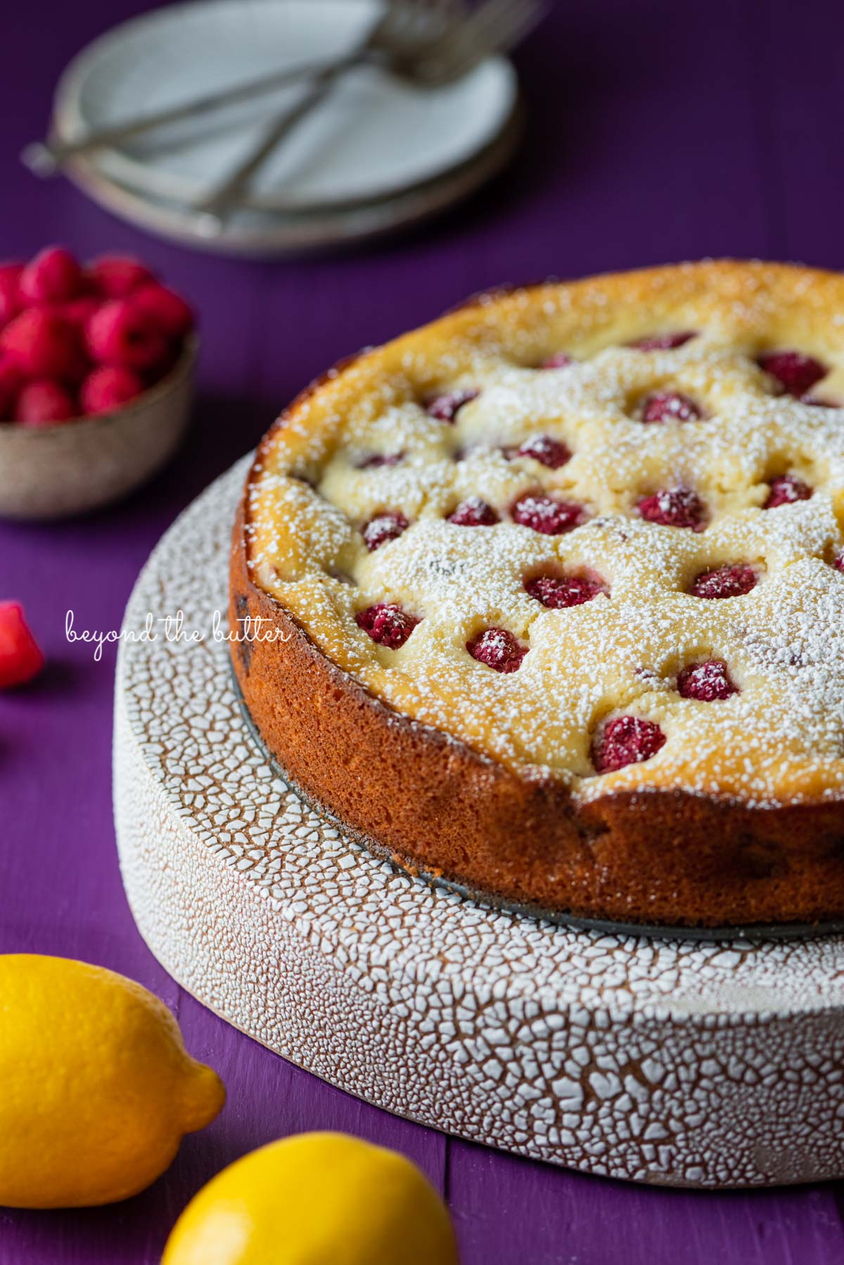 Lemon raspberry ricotta cake dusted with powdered sugar on crackled ceramic cake stand and purple wood background | © Beyond the Butter®