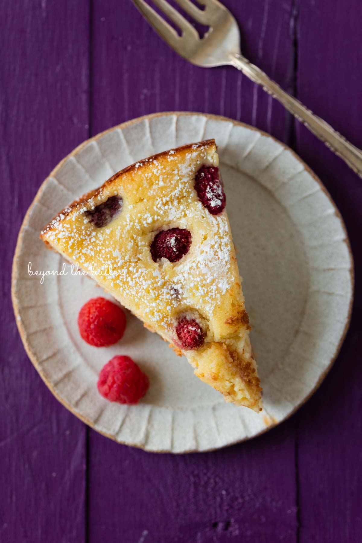 Slice of lemon raspberry ricotta cake dusted with powdered sugar on a small ceramic dessert plate and purple wood background | © Beyond the Butter®
