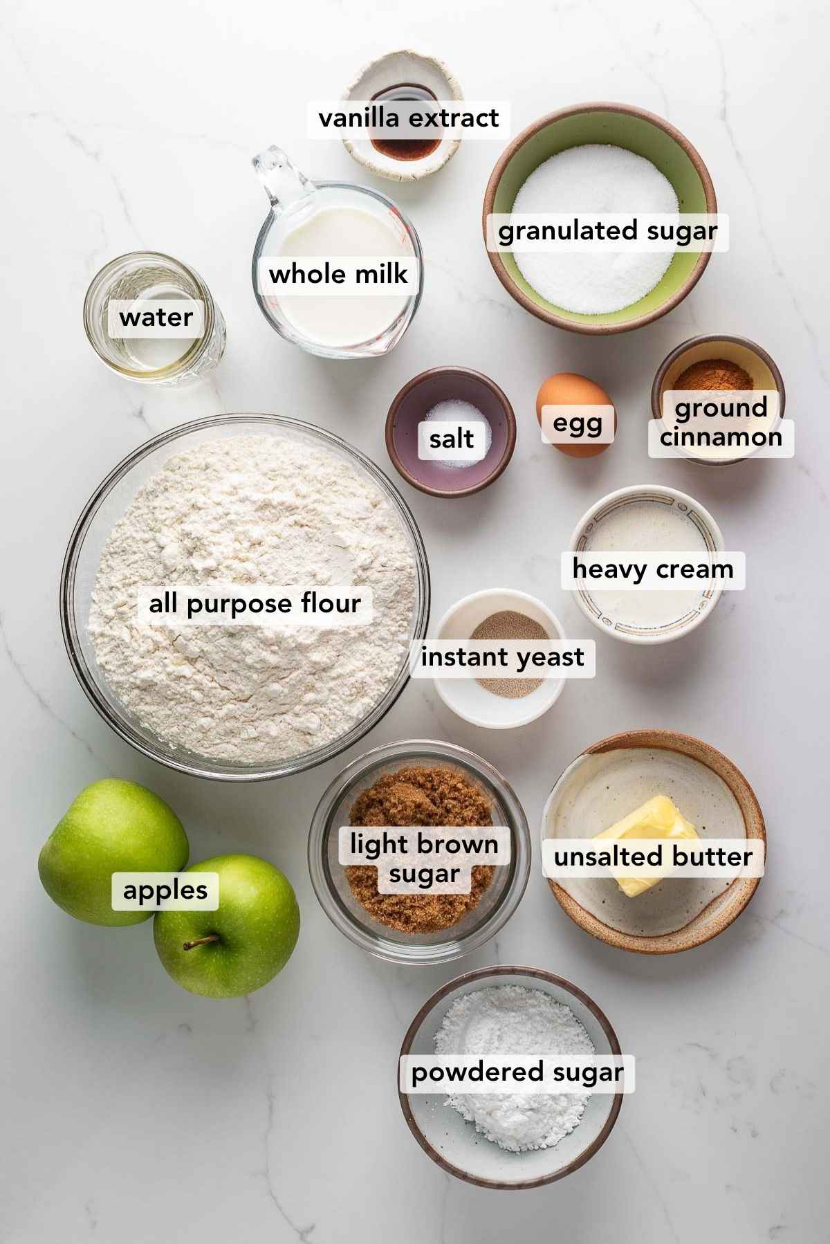 Ingredients in various size bowls labeled as vanilla extract, water, whole milk, granulated sugar, all purpose flour, salt, egg, instant yeast, heavy cream, ground cinnamon,apples, light brown sugar, butter, and powdered sugar on a white marbled background.