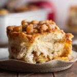 Apple streusel cinnamon roll on a ceramic dessert plate on a dark wood table with a pan of cinnamon rolls and apples in the background.