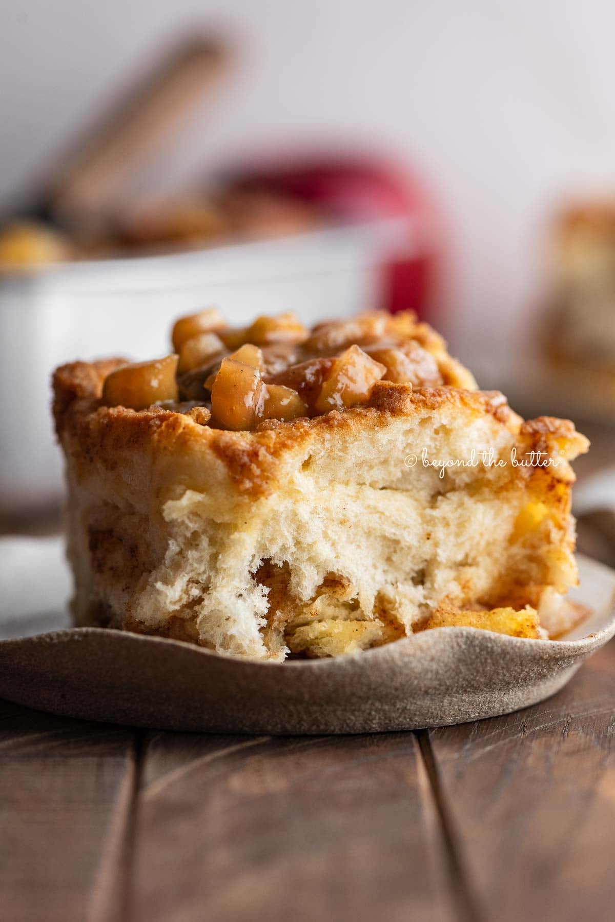 Apple streusel cinnamon roll on a ceramic dessert plate on a dark wood table with a pan of cinnamon rolls and apples in the background.