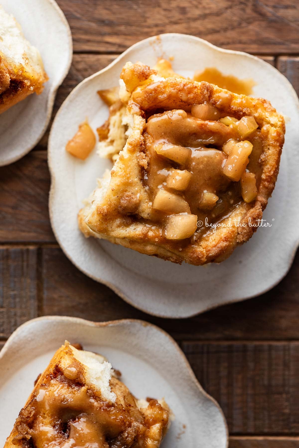 Apple cinnamon rolls topped with streusel and caramel apple glaze on ceramic dessert plates on a dark wood table.