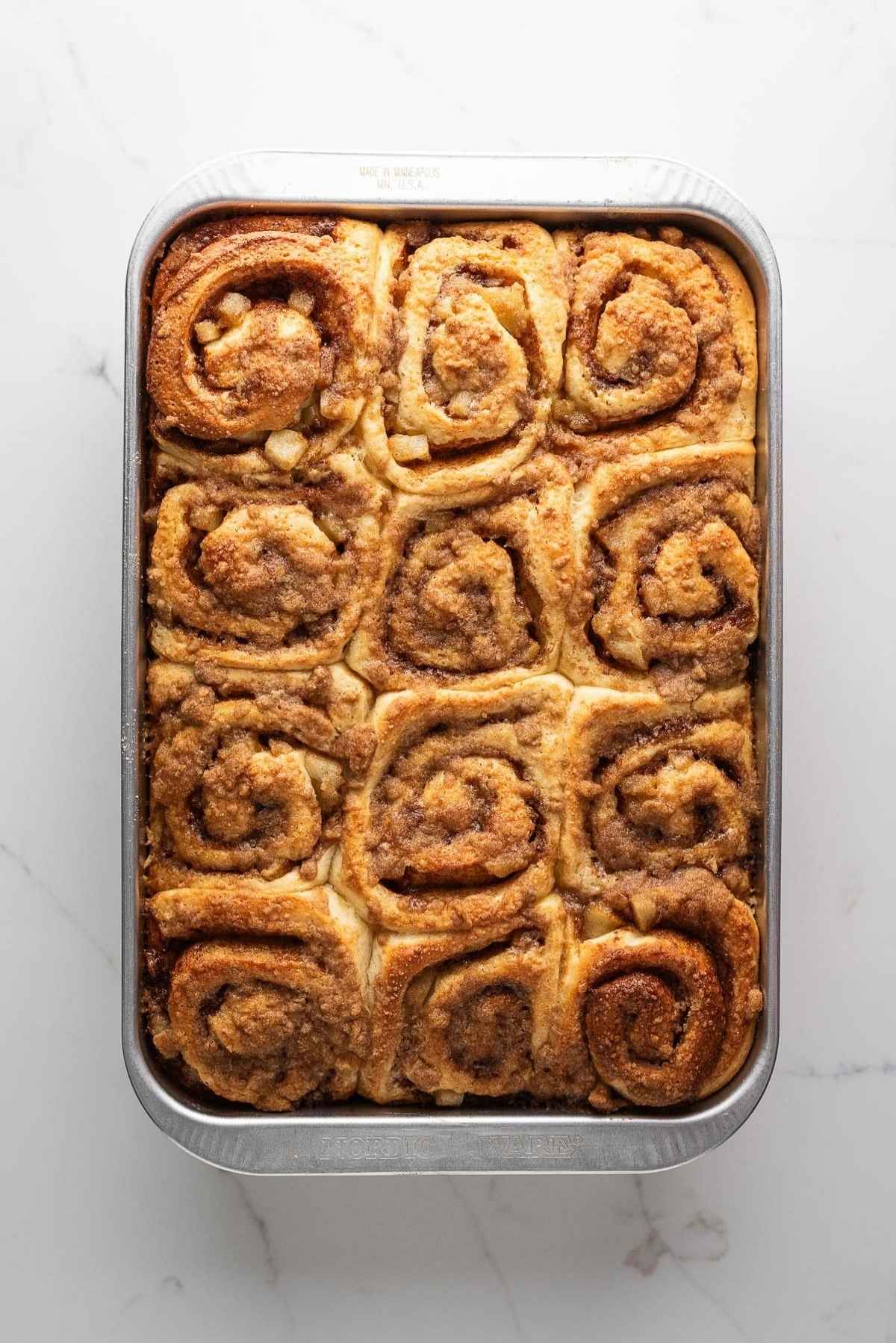 Baked apple cinnamon rolls topped with cinnamon streusel in a nine inch by thirteen inch metal baking pan on a light grey marbled background.