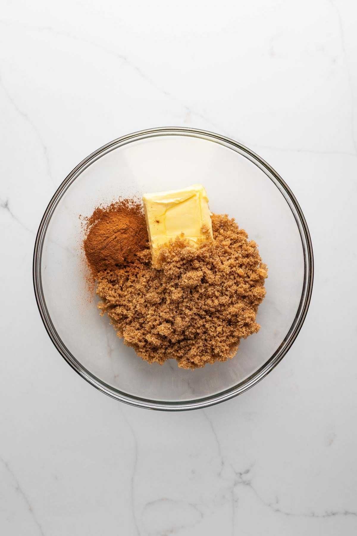 Brown sugar, butter, and cinnamon in a small glass mixing bowl on a light grey marbled background.