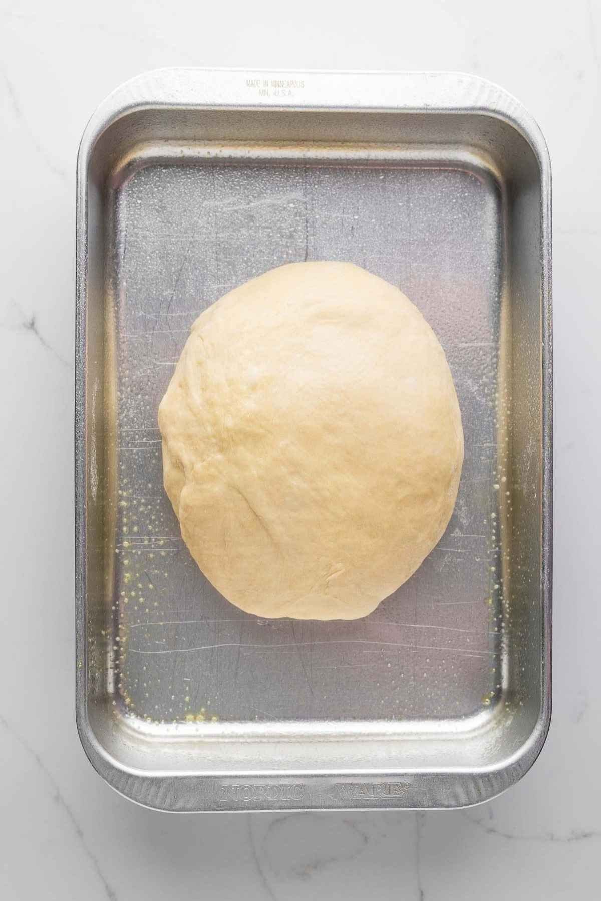 Dough placed in a greased nine inch by thirteen inch metal pan on a light grey marbled background.