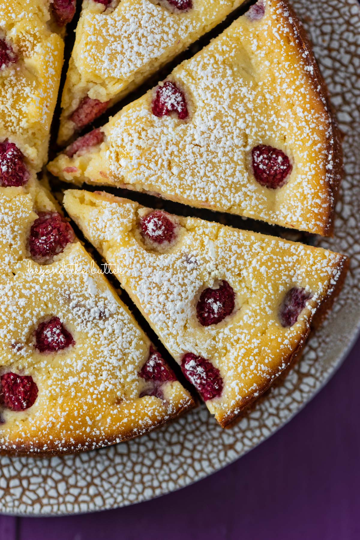 Sliced lemon raspberry ricotta cake on crackled ceramic cake stand and purple wood background | © Beyond the Butter®