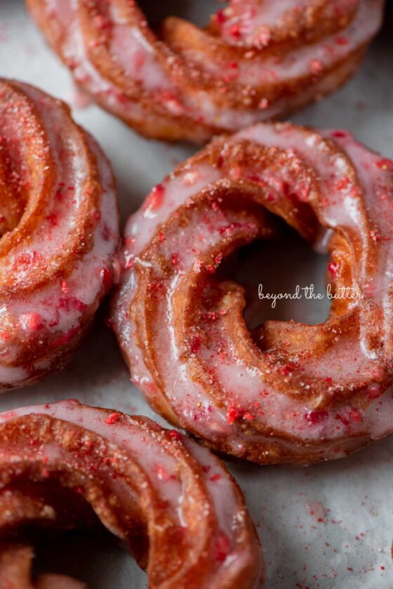 Strawberry glazed french crullers sprinkled with strawberry sugar on a parchment paper lined baking sheet | © Beyond the Butter®