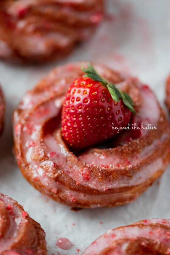 Strawberry glazed french crullers on a parchment paper lined baking sheet topped with a fresh strawberry.