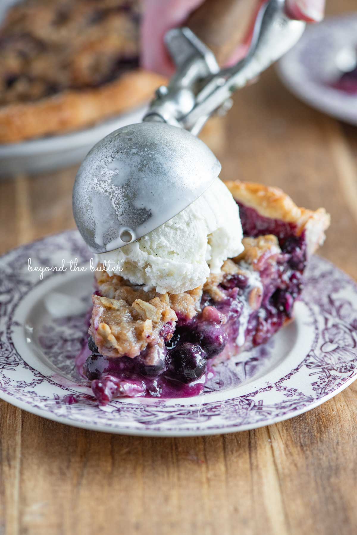 Slice of blueberry crumble pie with scoop of vanilla bean ice cream on top | © Beyond the Butter®
