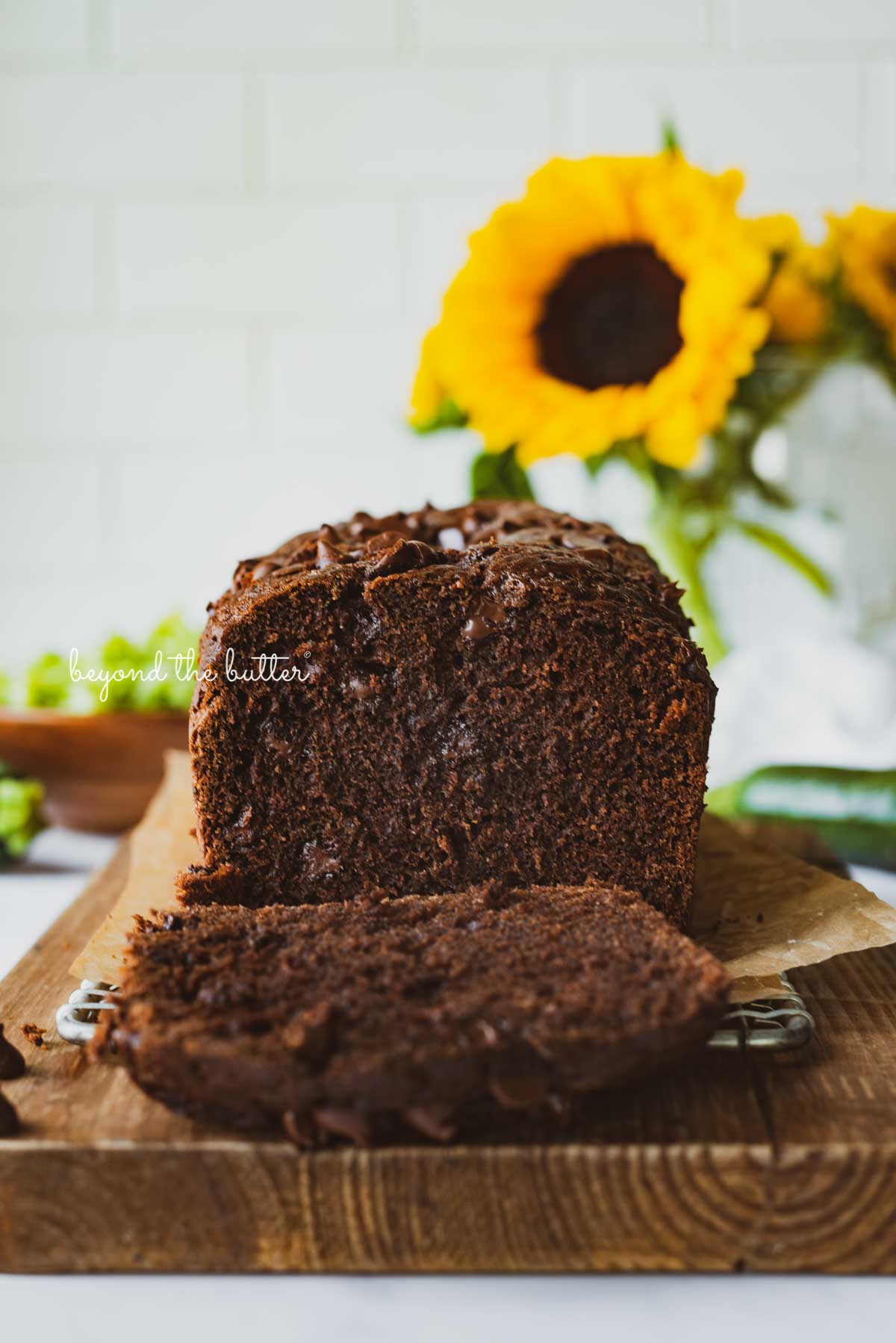Sliced chocolate zucchini bread on a cutting board with white tile background and sunflowers | © Beyond the Butter®