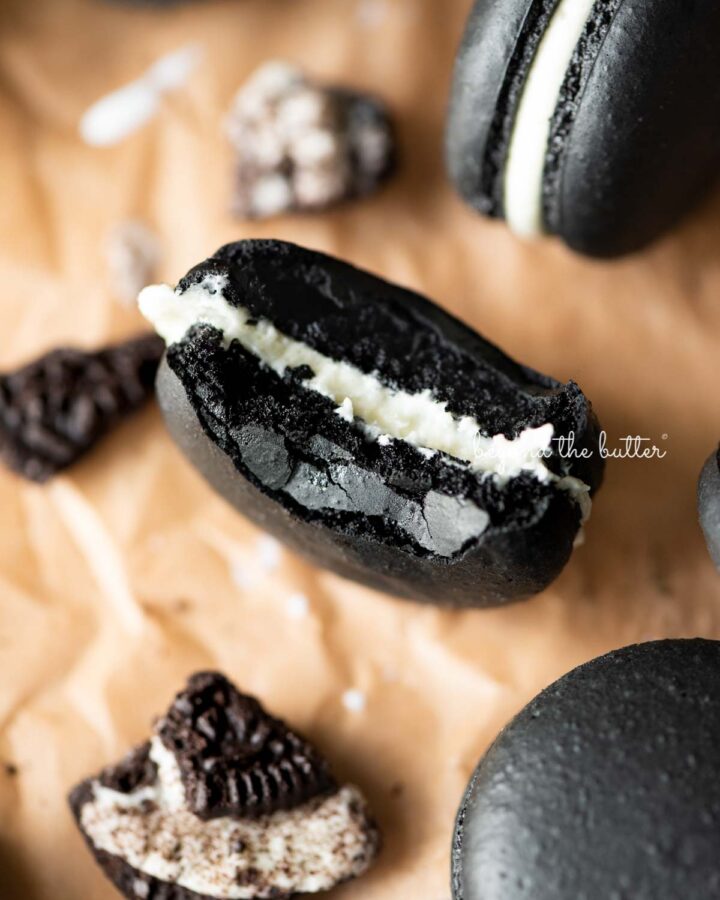 Half eaten oreo macaron on parchment paper surrounded by oreo macarons and broken pieces of oreo cookies.