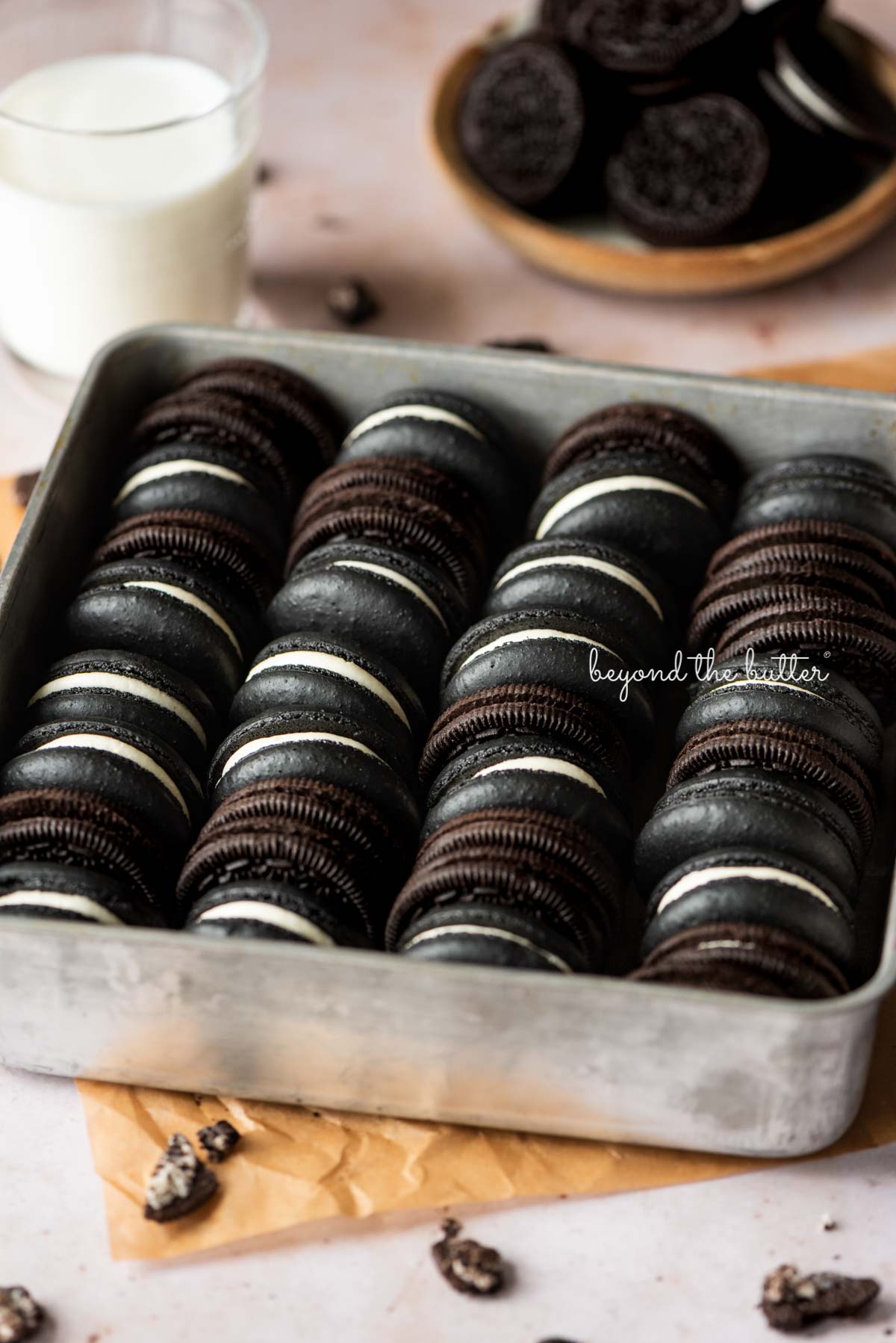 Oreo macarons and oreo cookies in a vintage baking tin on parchment paper with a glass of milk and bowl of oreo cookies in the background.