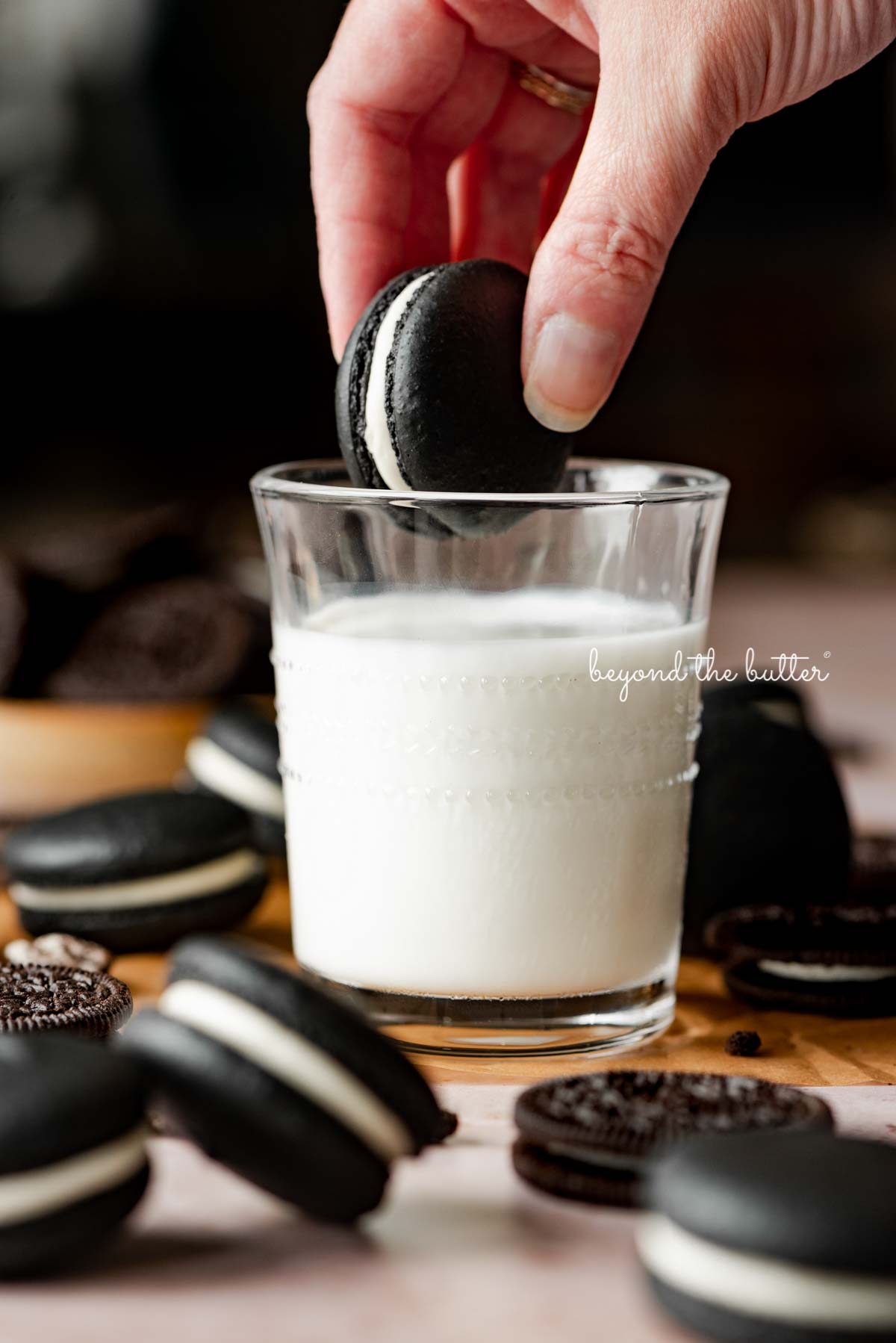 Hand dipping an oreo macaron in a glass of milk with more oreo macarons and oreo cookies around it | © Beyond the Butter®