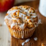 Apple cinnamon streusel muffin on a wood cutting board with slices of apples and vanilla glaze.