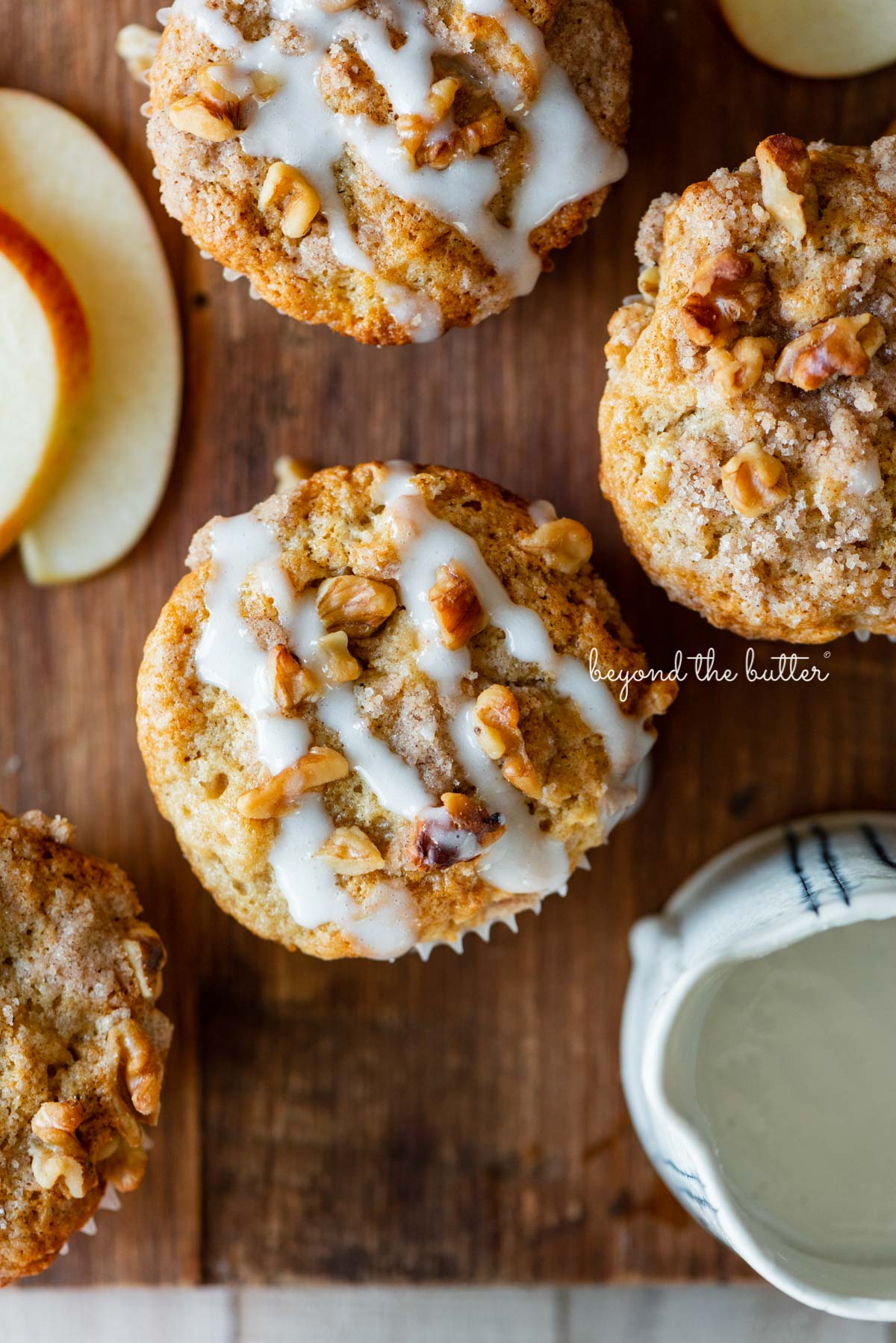Warm apple cinnamon streusel muffins on a wood cutting board with slices of apples and vanilla glaze | © Beyond the Butter®
