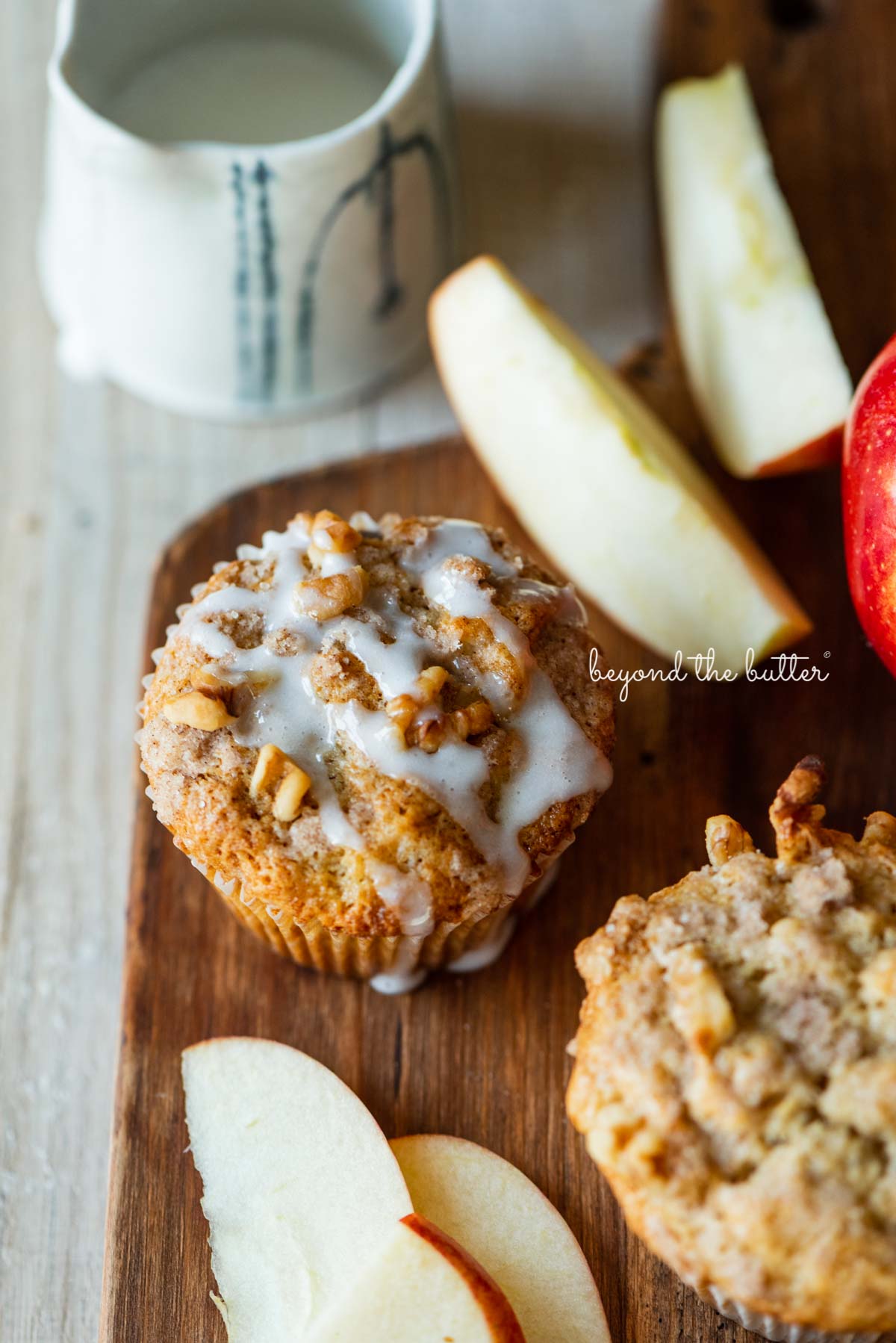 Warm apple cinnamon streusel muffins on a wood cutting board with slices of apples and vanilla glaze | © Beyond the Butter®
