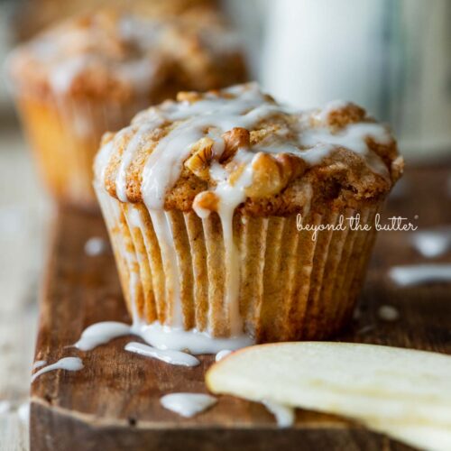 Warm apple cinnamon streusel muffins on a wood cutting board with slices of apples and vanilla glaze.