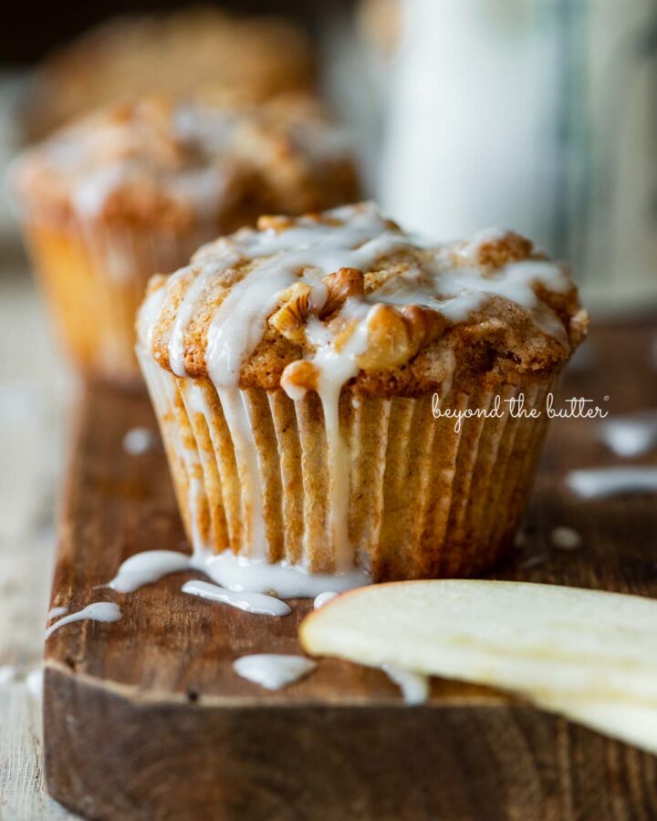 Warm apple cinnamon streusel muffins on a wood cutting board with slices of apples and vanilla glaze.