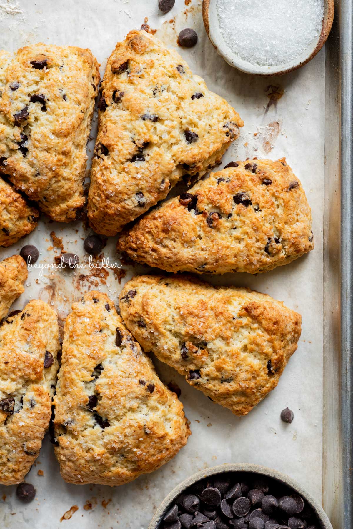 Easy chocolate chip scones on a parchment paper lined baking sheet with a bowl of chocolate chips and sparkling sugar | © Beyond the Butter®