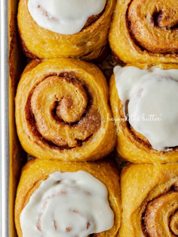 Six pumpkin cinnamon rolls with three topped with maple frosting placed on a small baking sheet.