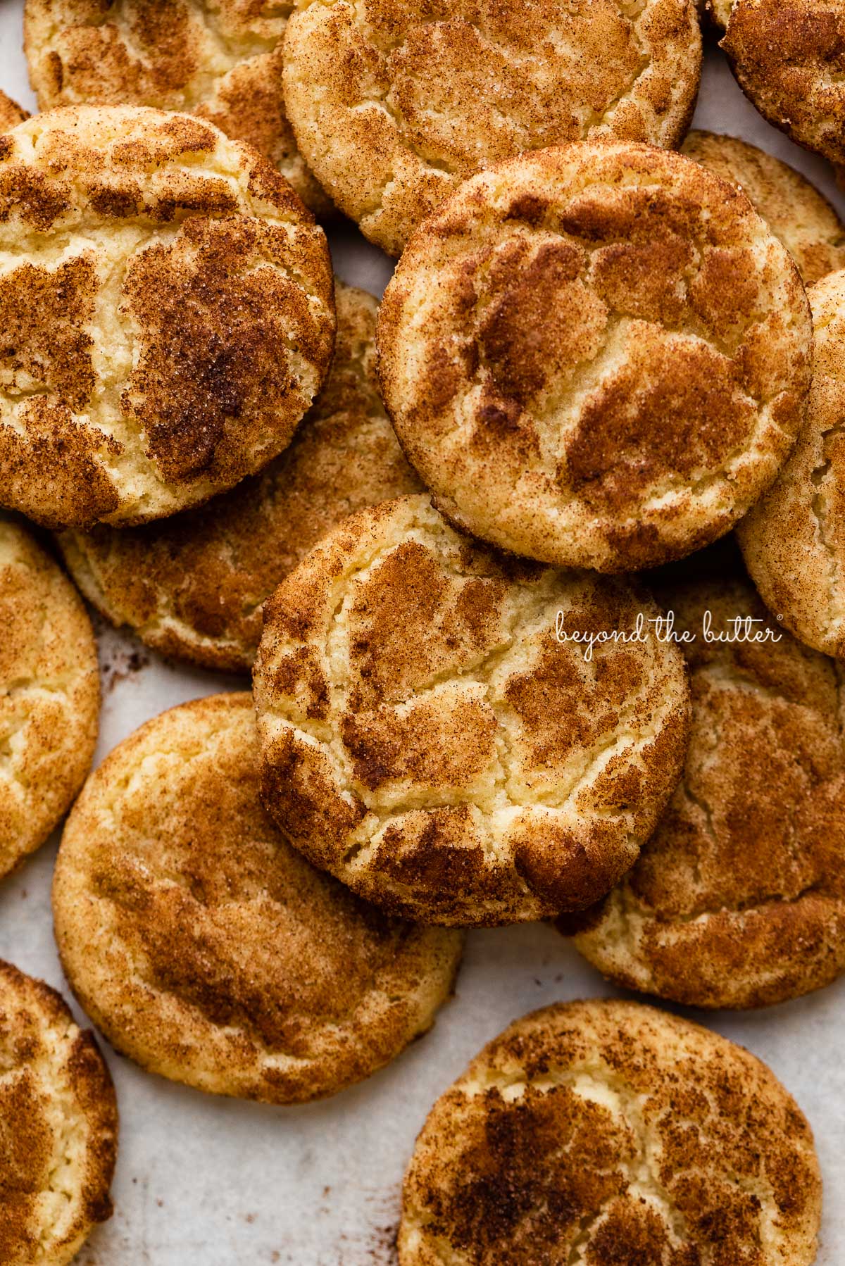 Piled snickerdoodle cookies on a parchment paper lined baking sheet | © Beyond the Butter®