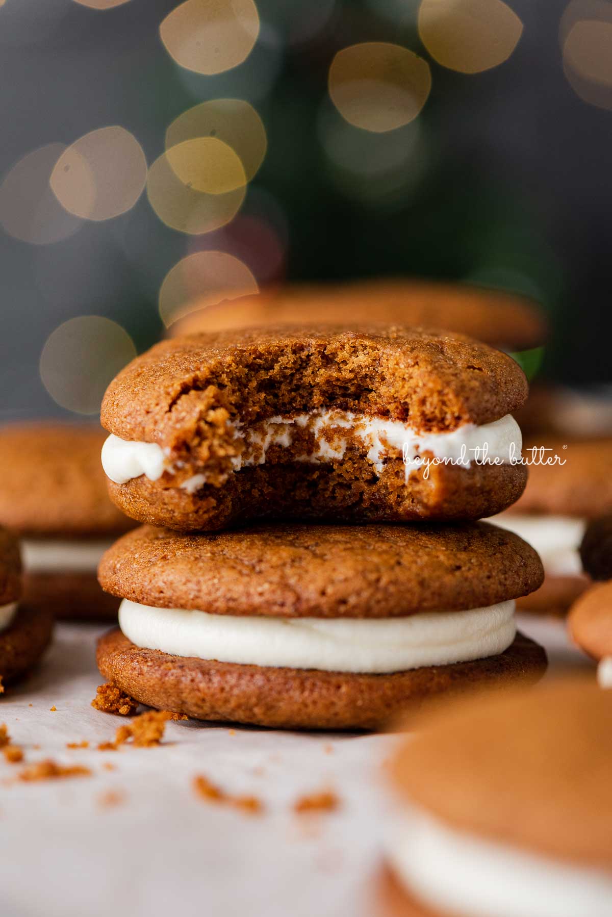 Half eaten gingerbread whoopie pie placed on top of another whoopie pie with a holiday tree background | © Beyond the Butter®