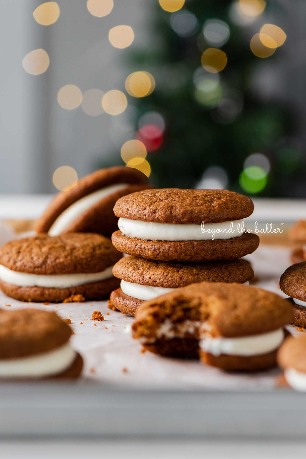 Stacked gingerbread whoopie pies on white parchment paper with holiday tree background | © Beyond the Butter®