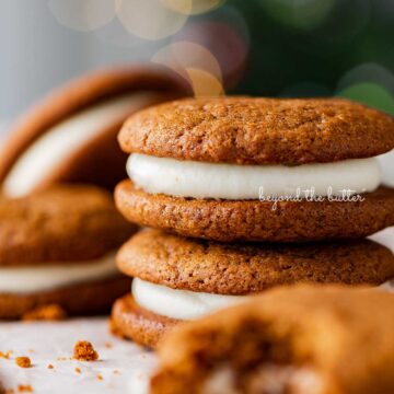 Stacked gingerbread whoopie pies on white parchment paper with holiday tree background | © Beyond the Butter®
