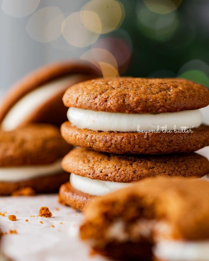 Stacked gingerbread whoopie pies on white parchment paper with holiday tree background.