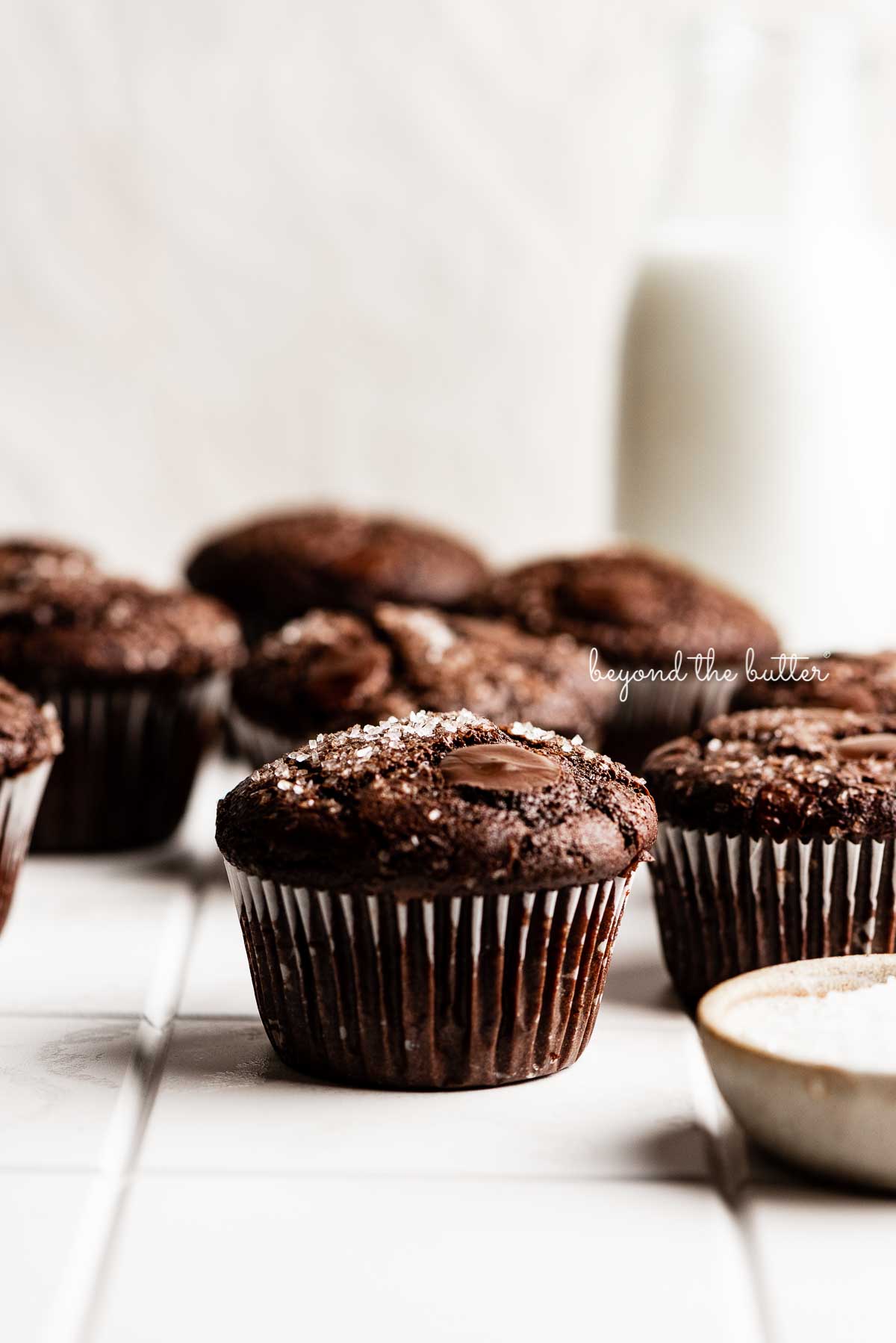 Double chocolate muffins on white tiled background with bottle of milk and small bowl of sparklin sugar to the side | © Beyond the Butter®