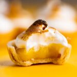 Mini lemon meringue pie with bite taken out surrounded by other mini pies on a yellow background.