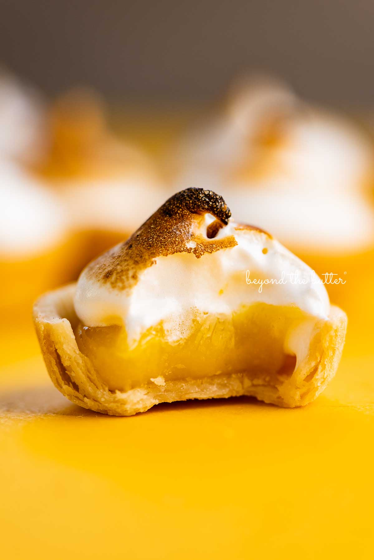 Mini lemon meringue pie with bite taken out surrounded by other mini pies on a yellow background | © Beyond the Butter@