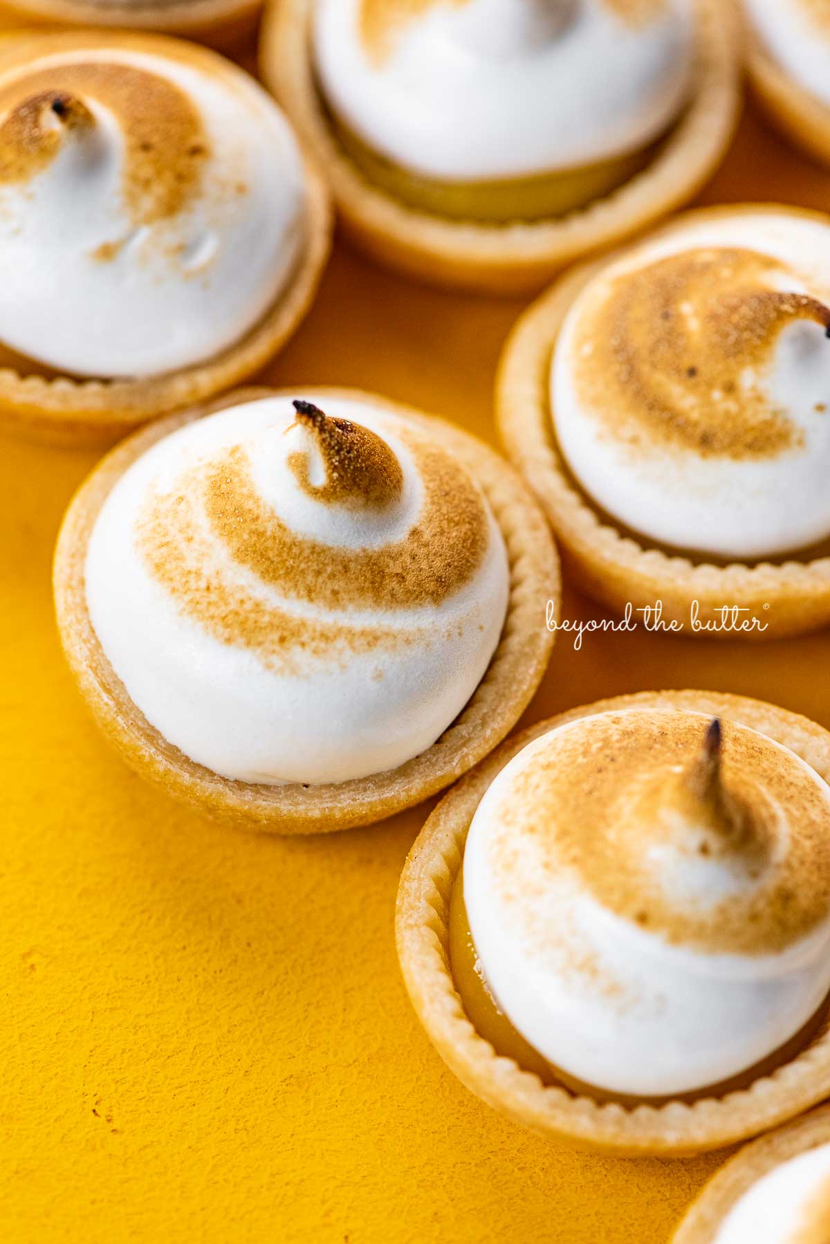 Angled image of mini lemon meringue pies on a yellow background | © Beyond the Butter®