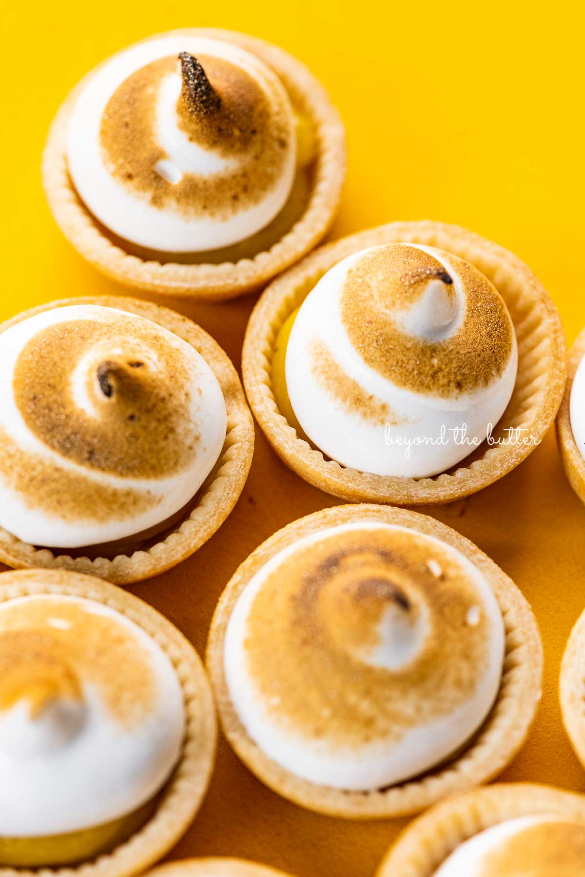 Angled image of mini lemon meringue pies on a yellow background | © Beyond the Butter®