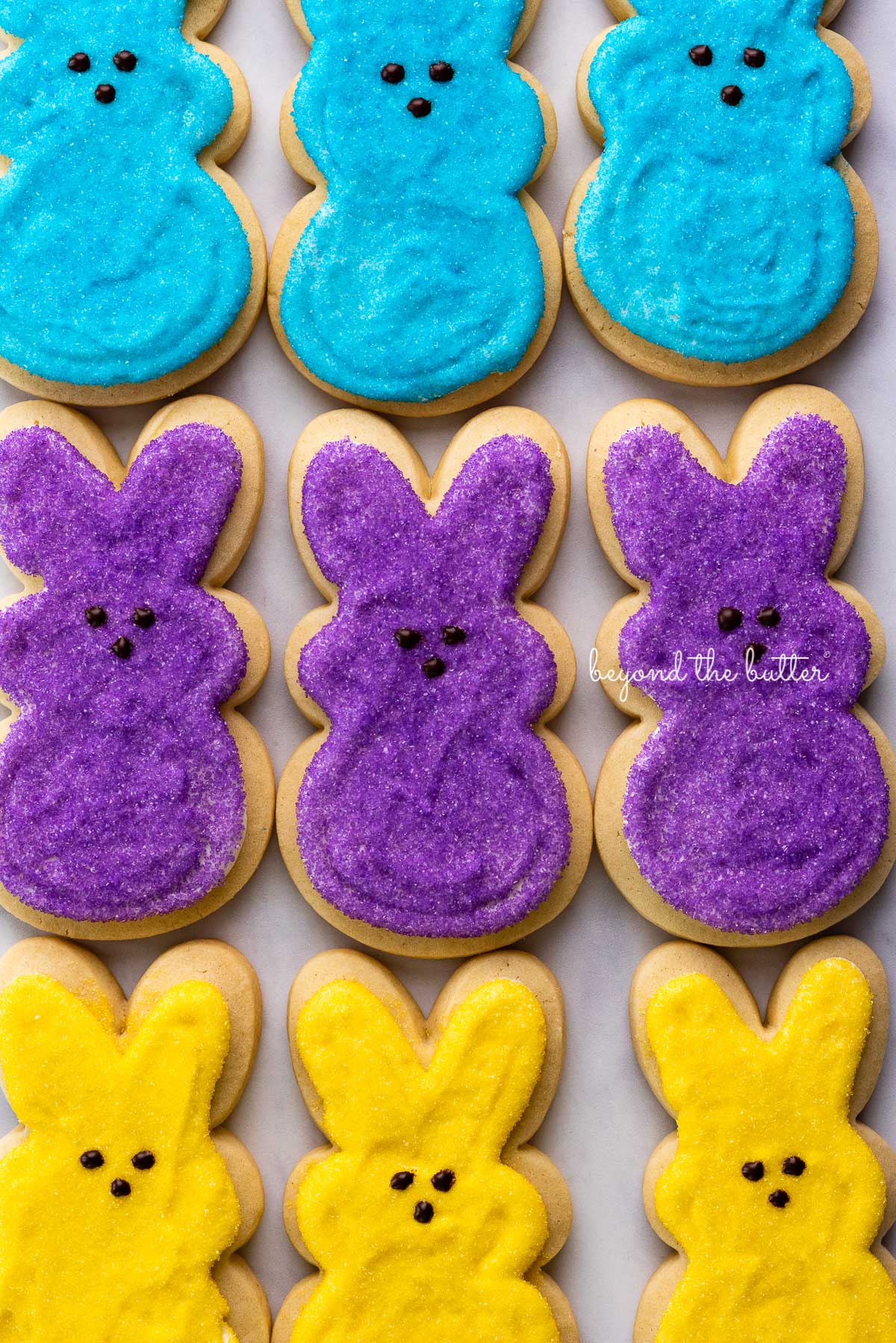 A row of blue, purple, and yellow easter bunny cut out sugar cookies on a marbled background | © Beyond the Butter®
