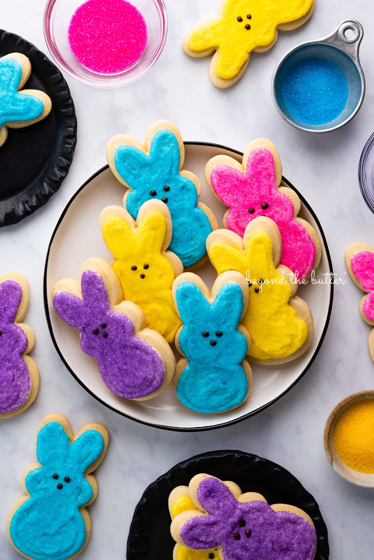 Plate full of Easter bunny cut out sugar cookies decorated in purple, pink, blue, and yellow sanding sugars with chocolate dots for eyes and noses on whie marbled background | © Beyond the Butter®