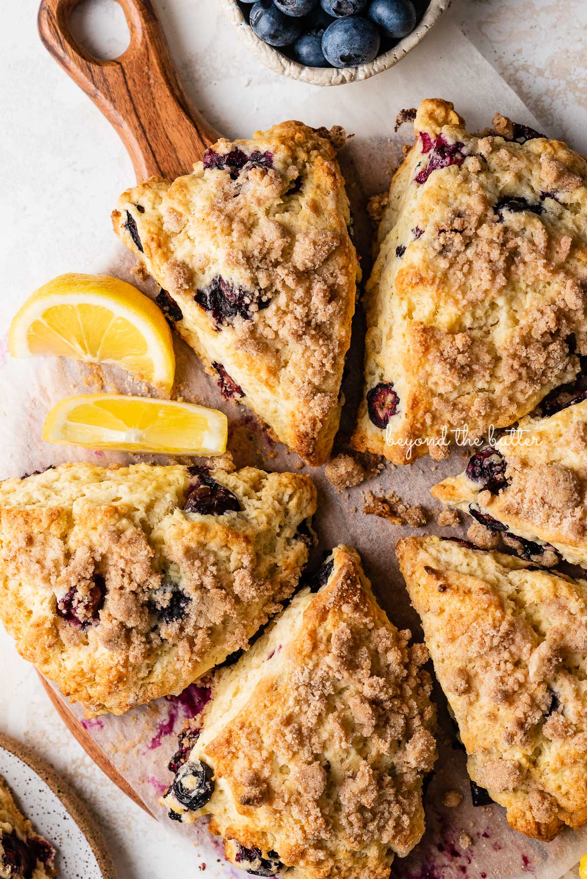 Lemon blueberry streusel scones surrounded by fresh blueberries on a parchment paper lined wood plate | © Beyond the Butter®