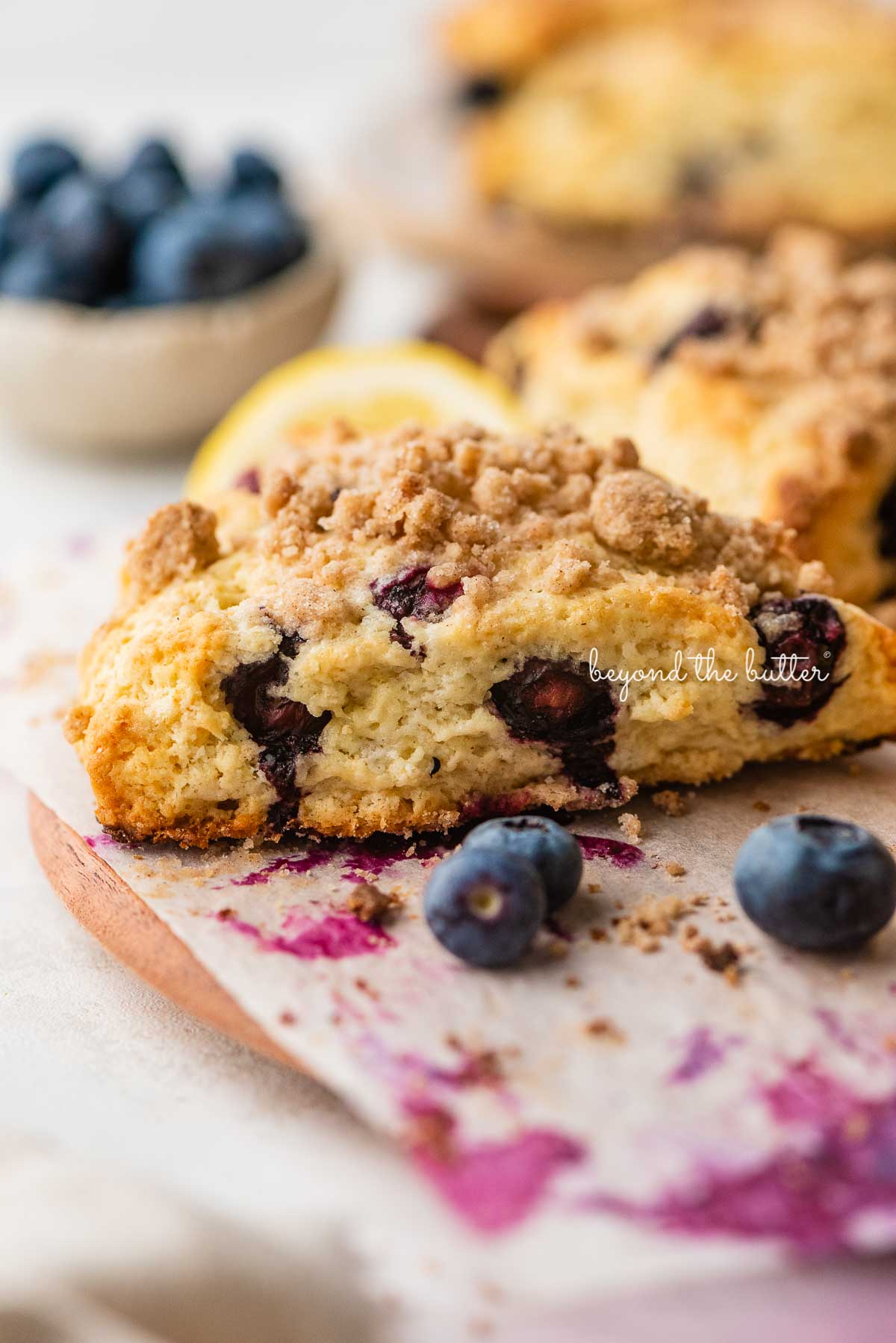 Lemon blueberry streusel scones surrounded by blueberries on a parchment paper lined wood plate | © Beyond the Butter®