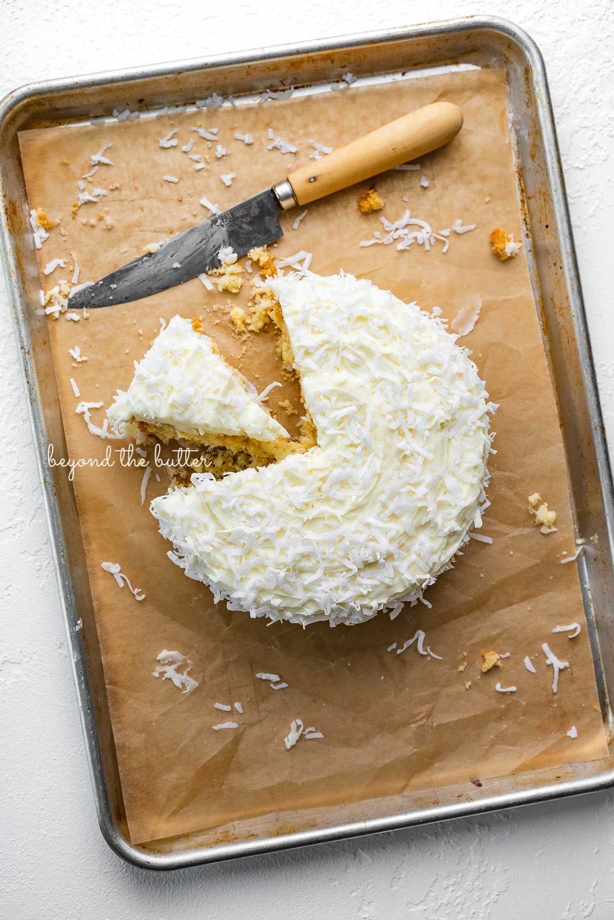 Single layer coconut cake frosted with coconut buttercream with slice cut out on natural parchment paper-lined baking sheet | © Beyond the Butter®