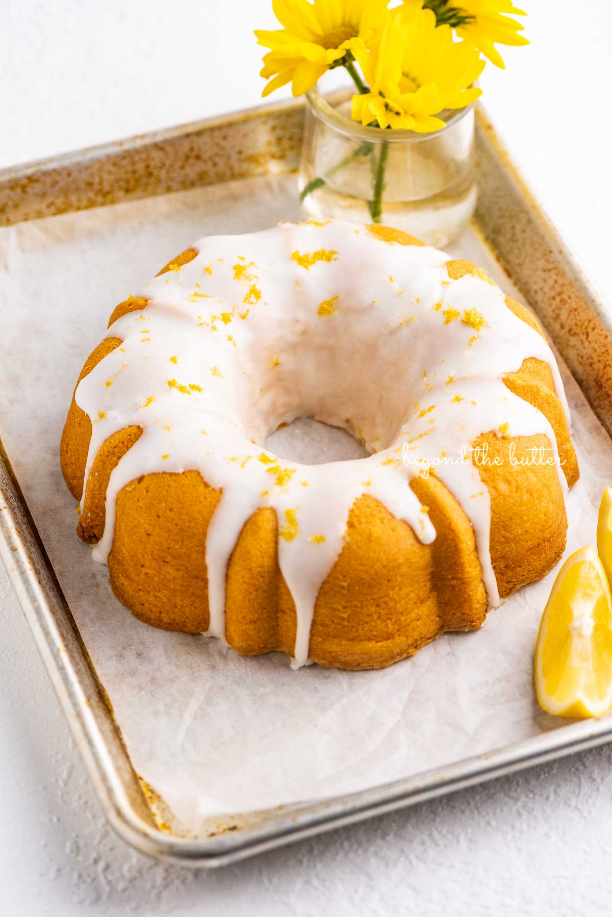 Small lemon cream cheese pound cake topped with a simple lemon glaze on a parchment paper lined baking sheet with sliced lemons and small bouquet of daisies | © Beyond the Butter®