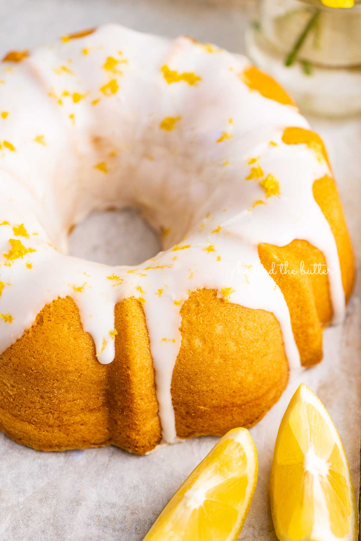 Small lemon cream cheese pound cake topped with a simple lemon glaze on a parchment paper lined baking sheet with sliced lemons | © Beyond the Butter®