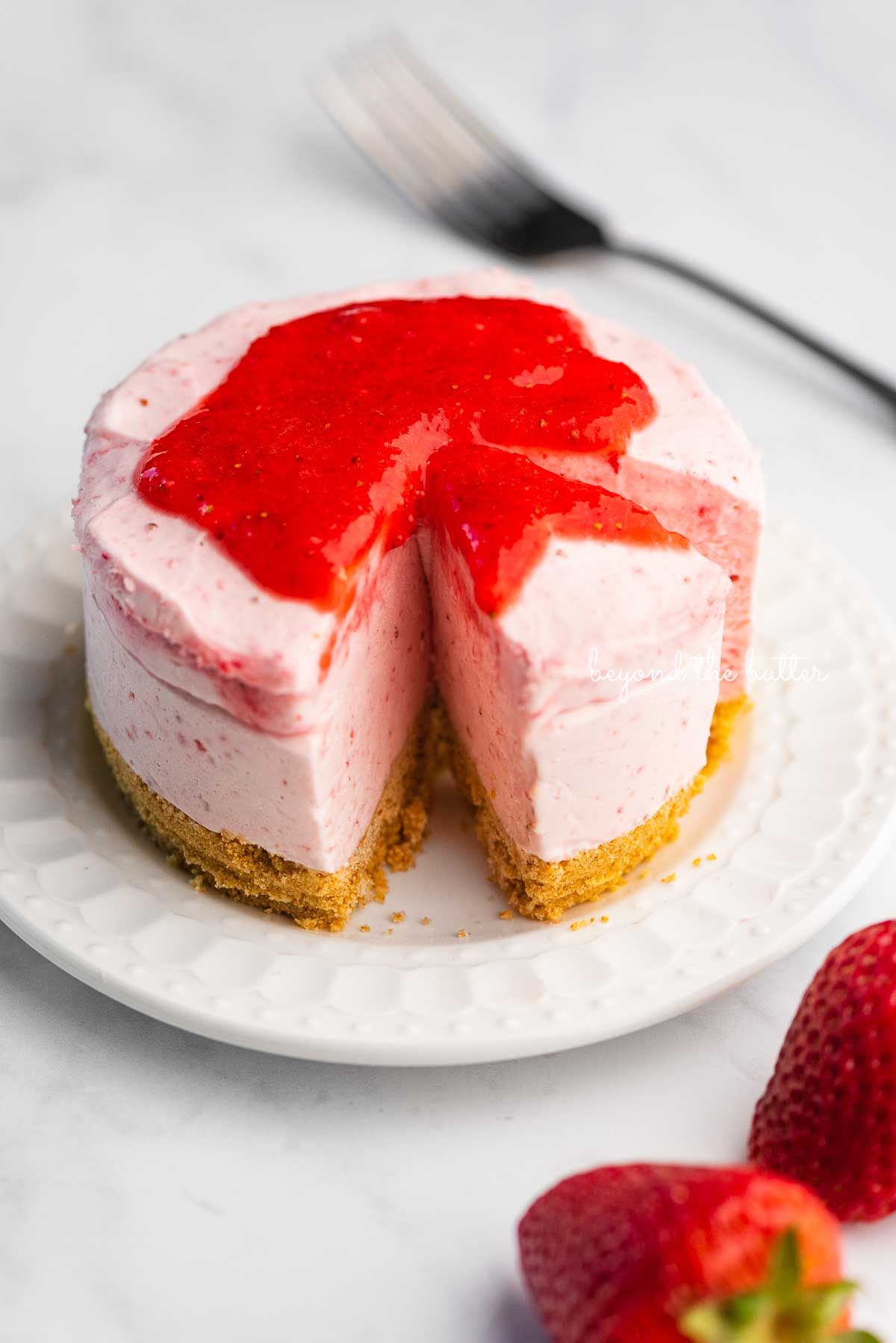 Small 4-inch no-bake strawberry cheesecake on a white dessert plate with a fork and fresh strawberries nearby on a marbled background | © Beyond the Butter®