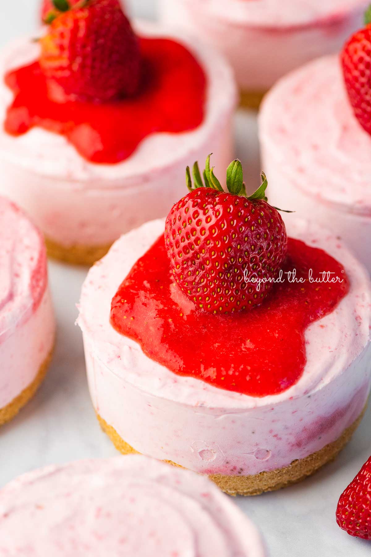 Individual no-bake strawberry cheesecakes topped with strawberry glaze and a fresh strawberry on a parchment paper-lined baking sheet.