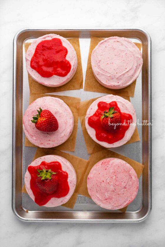 Individual no-bake strawberry cheesecakes topped with strawberry glaze and a fresh strawberry on a parchment paper-lined baking sheet | © Beyond the Butter®