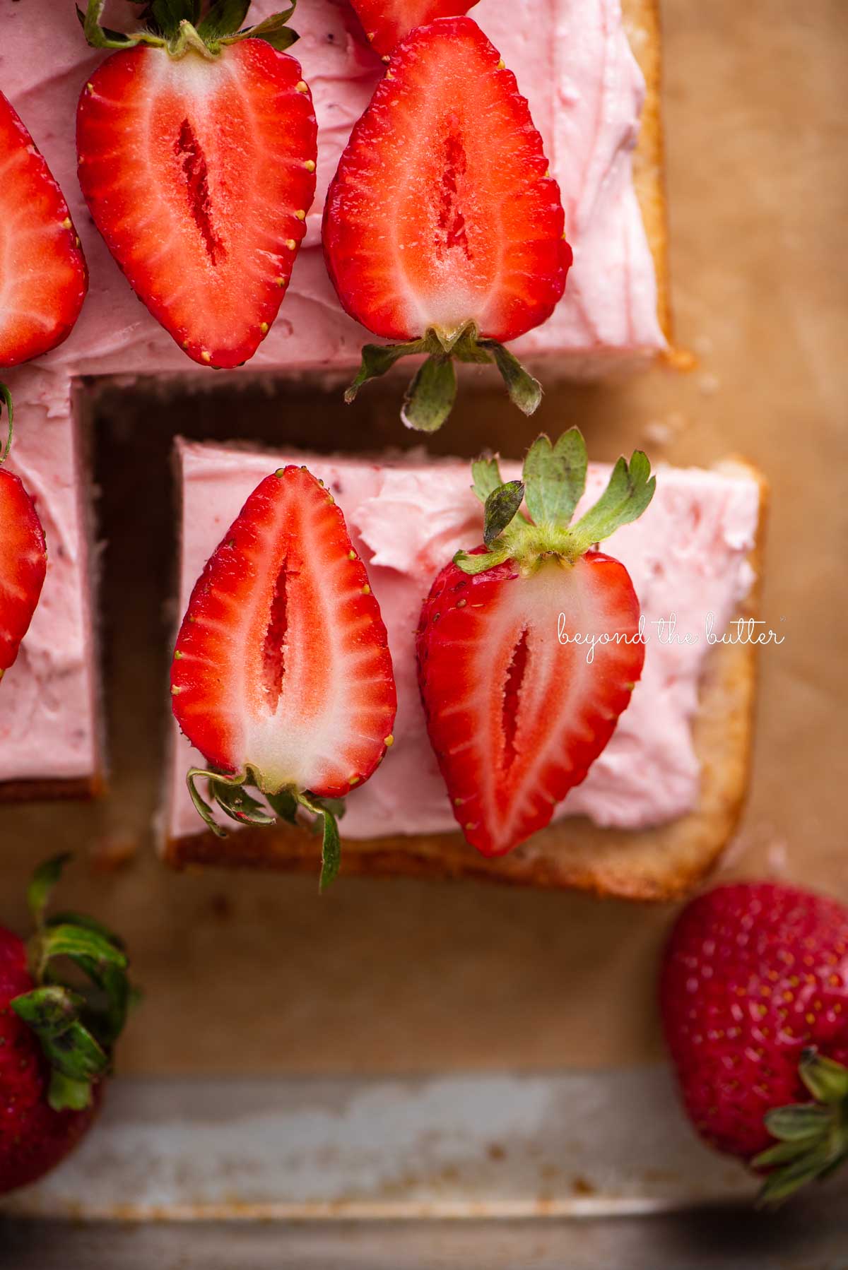 Piece of mini strawberry sheet cake topped with strawberry buttercream frosting and fresh sliced strawberries on parchment paper | © Beyond the Butter®